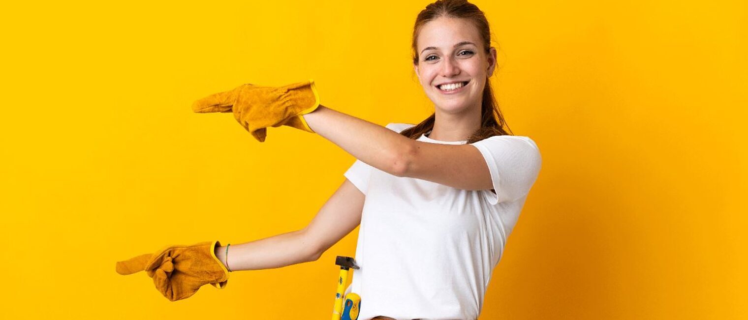 Mädchen mit Bauhandschuhen und Werkzeug um die Hüfte zeigt mit den Fingern nach links.