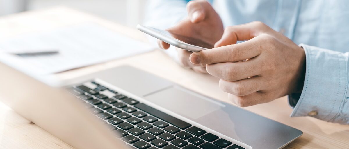 Männlicher Mitarbeiter scrollt oder schreibt einen Text über sein Smartphone. Vor ihm auf einem Holztisch steht ein Laptop. Young male employee scrolling or texting in smartphone over laptop keypad while sitting by wooden table Schlagwort(e): Adult, Business, Business Person, Businessman, Busy, Caucasian Ethnicity, Close-up, Communication, Computer, Computer Keyboard, Connection, Data, Desk, E-Mail, E-commerce, Equipment, Finance, Global Communications, Holding, Human Finger, Human Hand, Indoors, Internet, Laptop, Lifestyles, Males, Men, Mobile Phone, Mobility, Modern, Occupation, Office, One Person, PC, Part Of, People, Place of Work, Planning, Professional Occupation, Sitting, Table, Technology, Touching, Typing, Unrecognizable Person, Using Computer, Using Laptop, Wireless Technology, Working