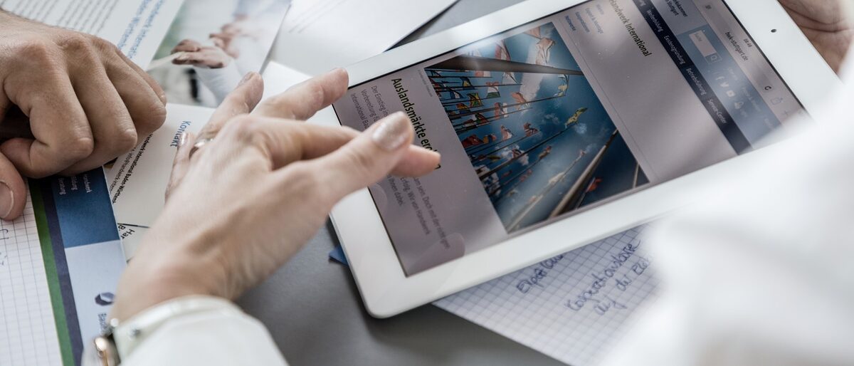 Frau zeigt auf einem Tablet einem Kunden die Webseite von Handwerk International BW. Auf dem Tisch liegen mehrere Broschüren verteilt. Handwerk International Baden-Württemberg, Enterprise Europe Net