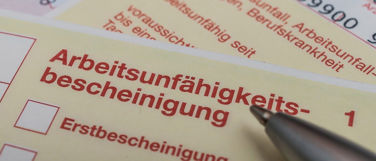 Silberner Kugelschreiber zeigt auf Arbeitsunfähigkeitsbescheinigung. arbeitsunfähigkeitsbescheinigung; krankenschein; arbeitsunfähig; krankgeschrieben; erwerbsunfähigkeit; kassenpatient; ansteckungsgefahr; krank geschrieben; krankengeld; krankenkasse; arbeitgeber; fehlzeit; arzt; arztbesuch; attest; bescheinigung; blau; einnahme; einnehmen; erkrankung; erwerbsunfähig; euro; fehlzeiten; formular; gesundheitskarte; grippewelle; hausarzt; infekt; infektion; krank; krankenversicherung; krankheit; krankheitsfall; krankmeldung; krankschreiben; krankschreibung; kugelschreiber; lohnfortzahlung; machen; patient; schein; schreibtisch; stift; tablette; tabletten; tastatur; untersuchen; untersuchung; vordruck; vorlage;