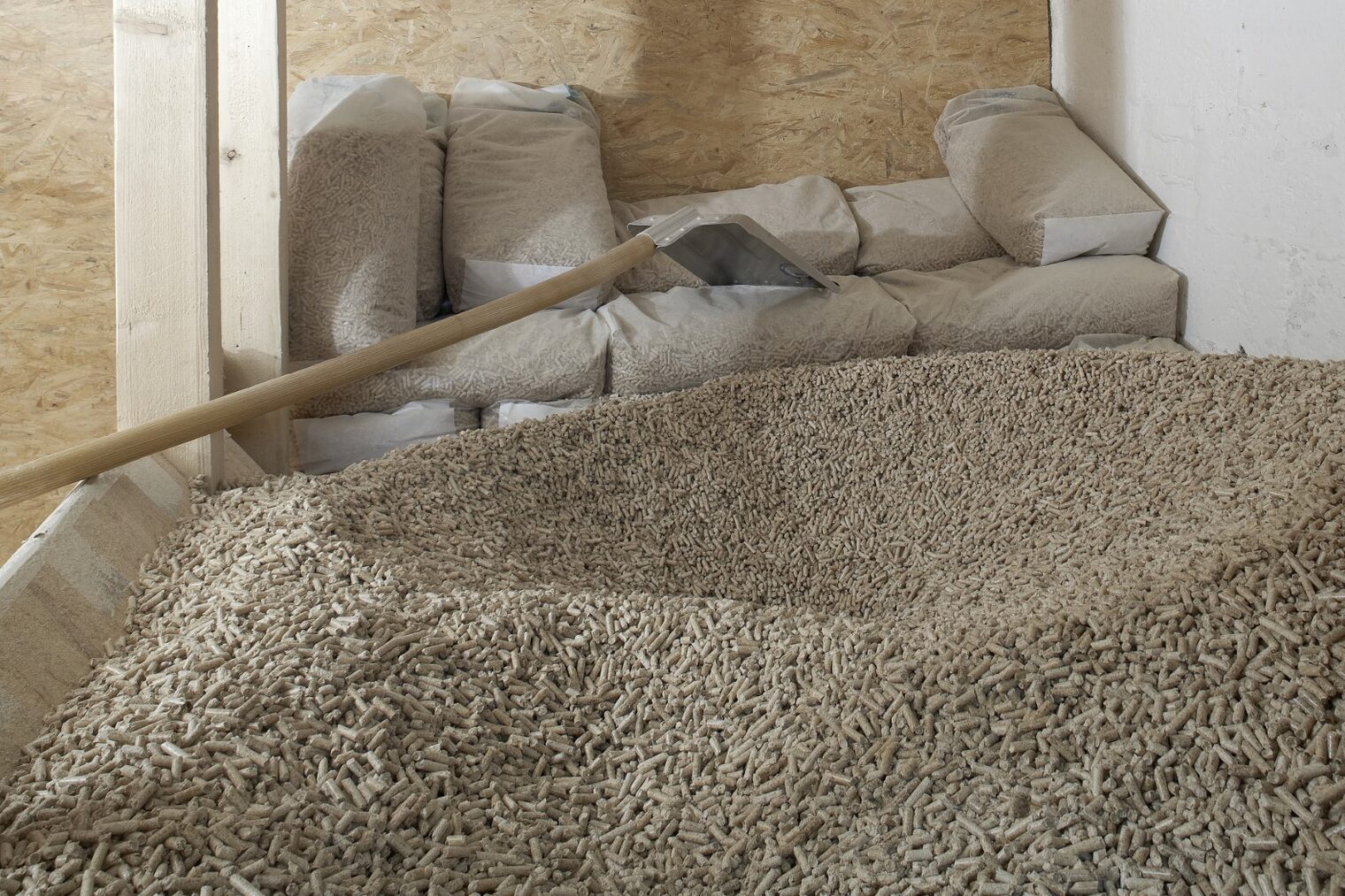 Vorne ein großer Haufen Holzpellets, dahinter eine Schaufel und abgepackte Beutel mit Holzpellets als Inhalt.