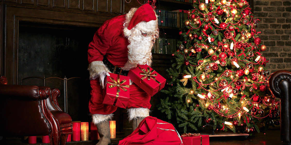 Weihnachtsmann mit Geschenken in der Hand. Neben ihm steht ein Weihnachtsbaum und hinter ihm ist ein Kamin. Weihnachtsmotiv 2021