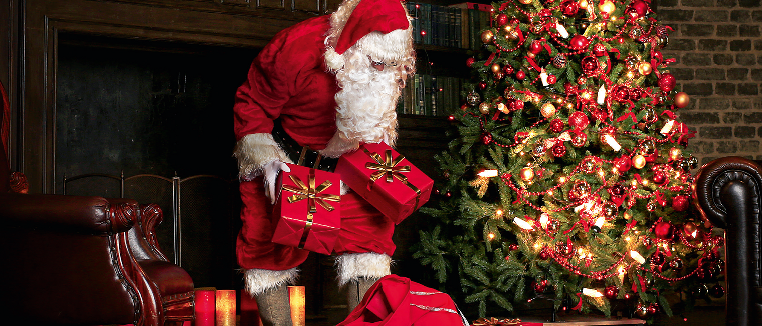 Weihnachtsmann mit Geschenken in der Hand. Neben ihm steht ein Weihnachtsbaum und hinter ihm ist ein Kamin. Weihnachtsmotiv 2021