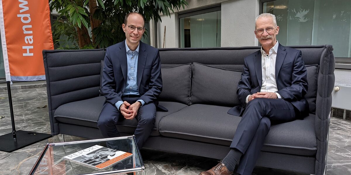 Walter Bantleon und Gerd Lutz sitzen auf einer Couch. Im Hintergrund steht eine Fahne vor ihnen ein kleiner Glastisch.