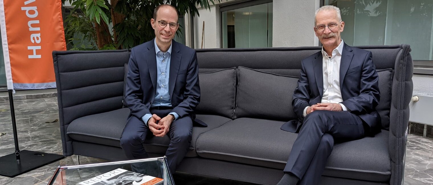 Walter Bantleon und Gerd Lutz sitzen auf einer Couch. Im Hintergrund steht eine Fahne vor ihnen ein kleiner Glastisch.