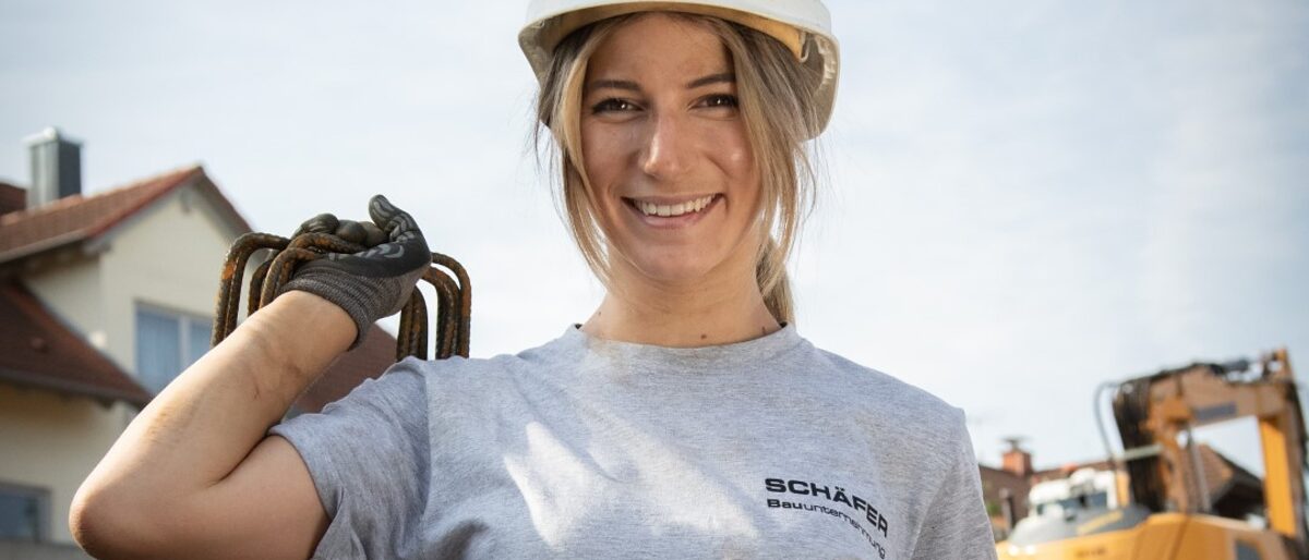 Maurermeisterin Julia Schäfer mit Bauhelm und Werkzeug in der Hand. Im Hintergrund ein Bagger und ein Haus.