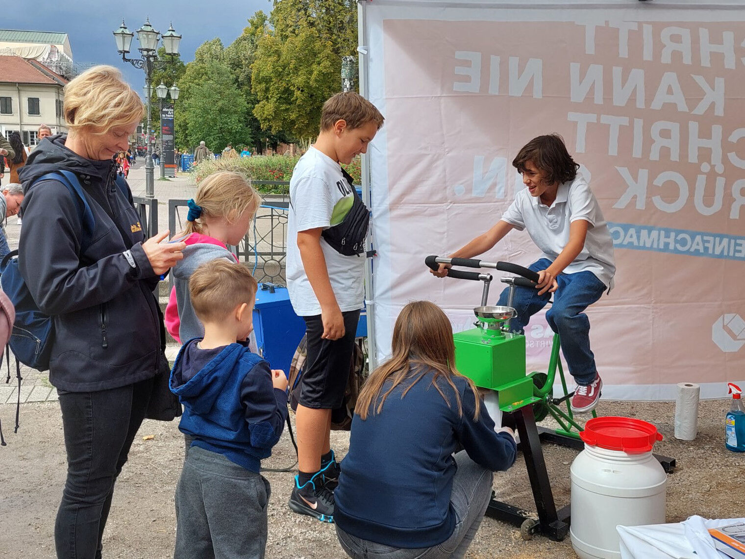 Jugendlicher tritt beim Müslifahrrad in die Pedale. Links davon steht eine Familie und wartet.