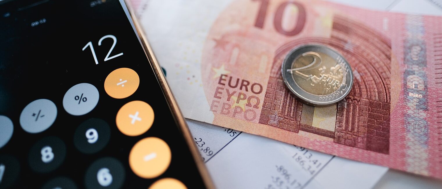 Linke Seite ein Taschenrechner auf Smartphone mit der Zahl 12 und auf der linken Seite ein 10 Euro-Schein und eine 2 Euro Münze daraufliegend.