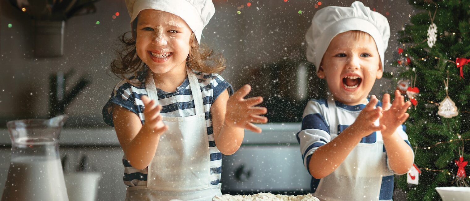 Zwei Kinder backen Weihnachtsgebäck und freuen sich. Im Hintergund ein Weihnachtsbaum in einer Backstube.