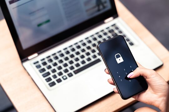 Hand mit Smartphone und einem Schloss-Symbol auf dem Bildschirm. Dahinter Laptop auf einem Schreibtisch.
